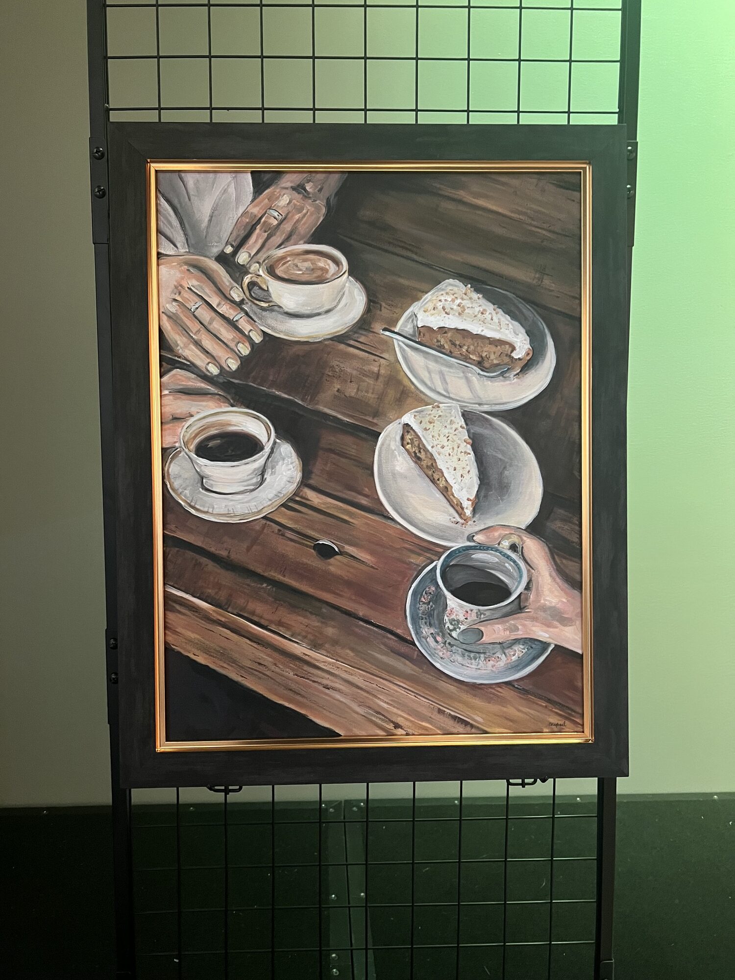Perspectives, Nicole’s acrylic painting of hands around coffee cups and slices of carrot cake on a wooden table, in a dark frame with a gold edge