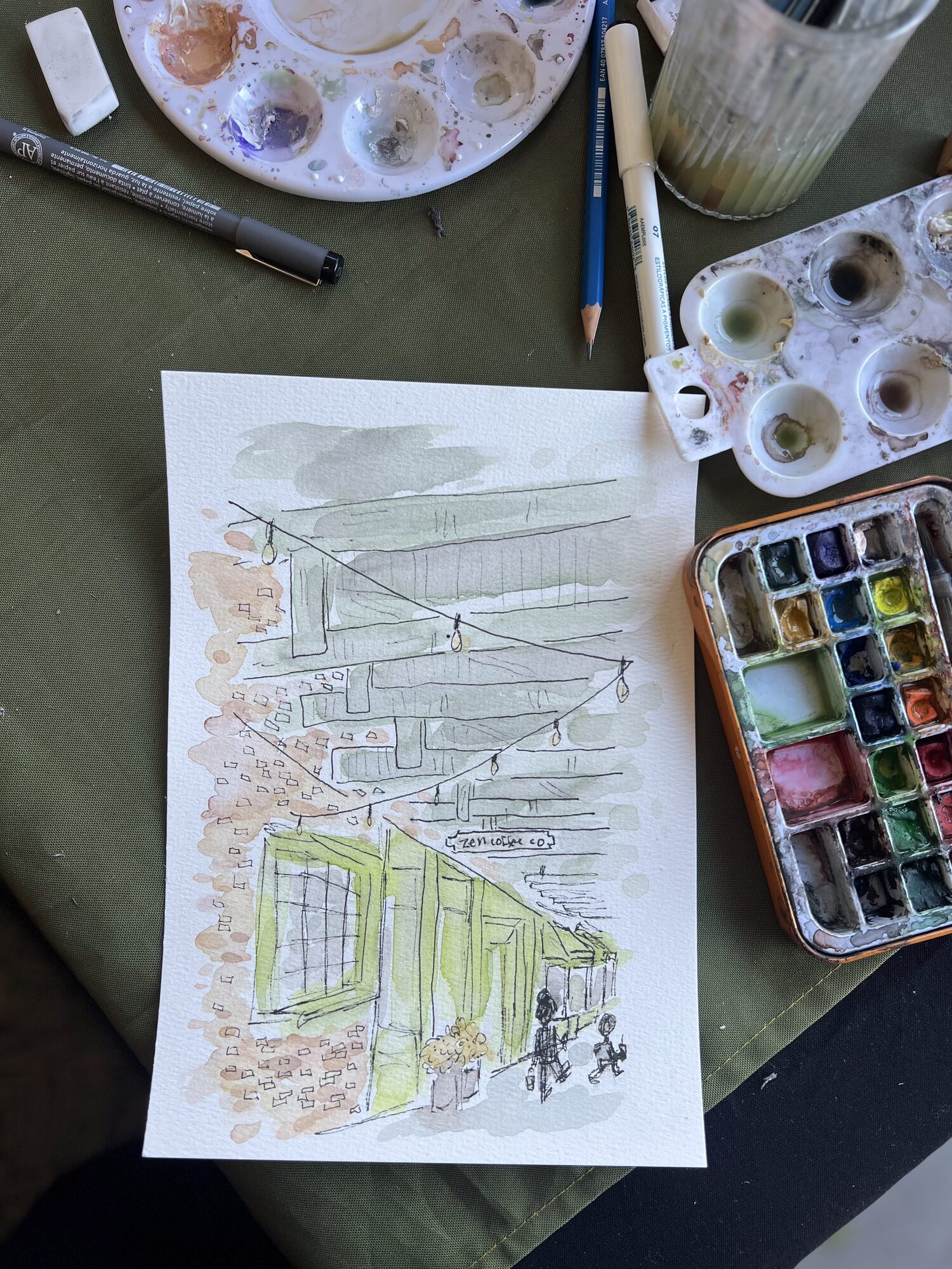 Nicole’s ink and watercolor painting of Zen Coffee’s brick front with its yellow-green window frames and string lights, beside her paint palette