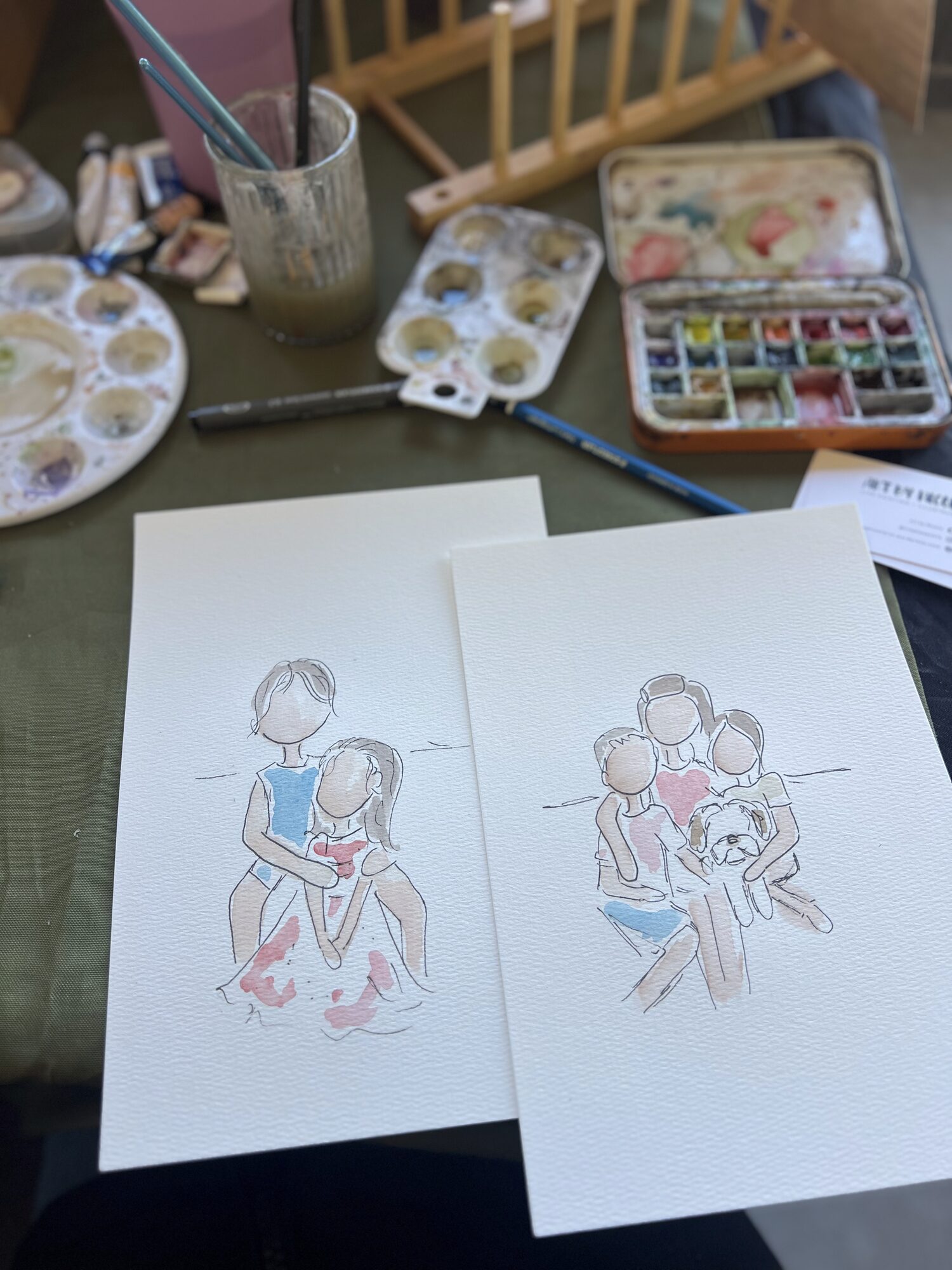 Two watercolor mini portraits from the pop-up on Nicole’s table, beside her palettes and brushes