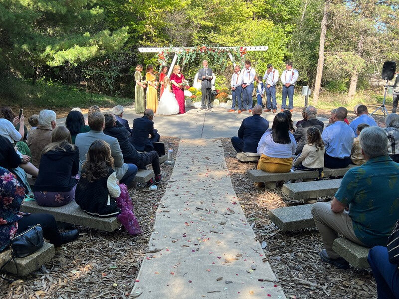 A wooded outdoor wedding ceremony, guests seated on either side of the aisle