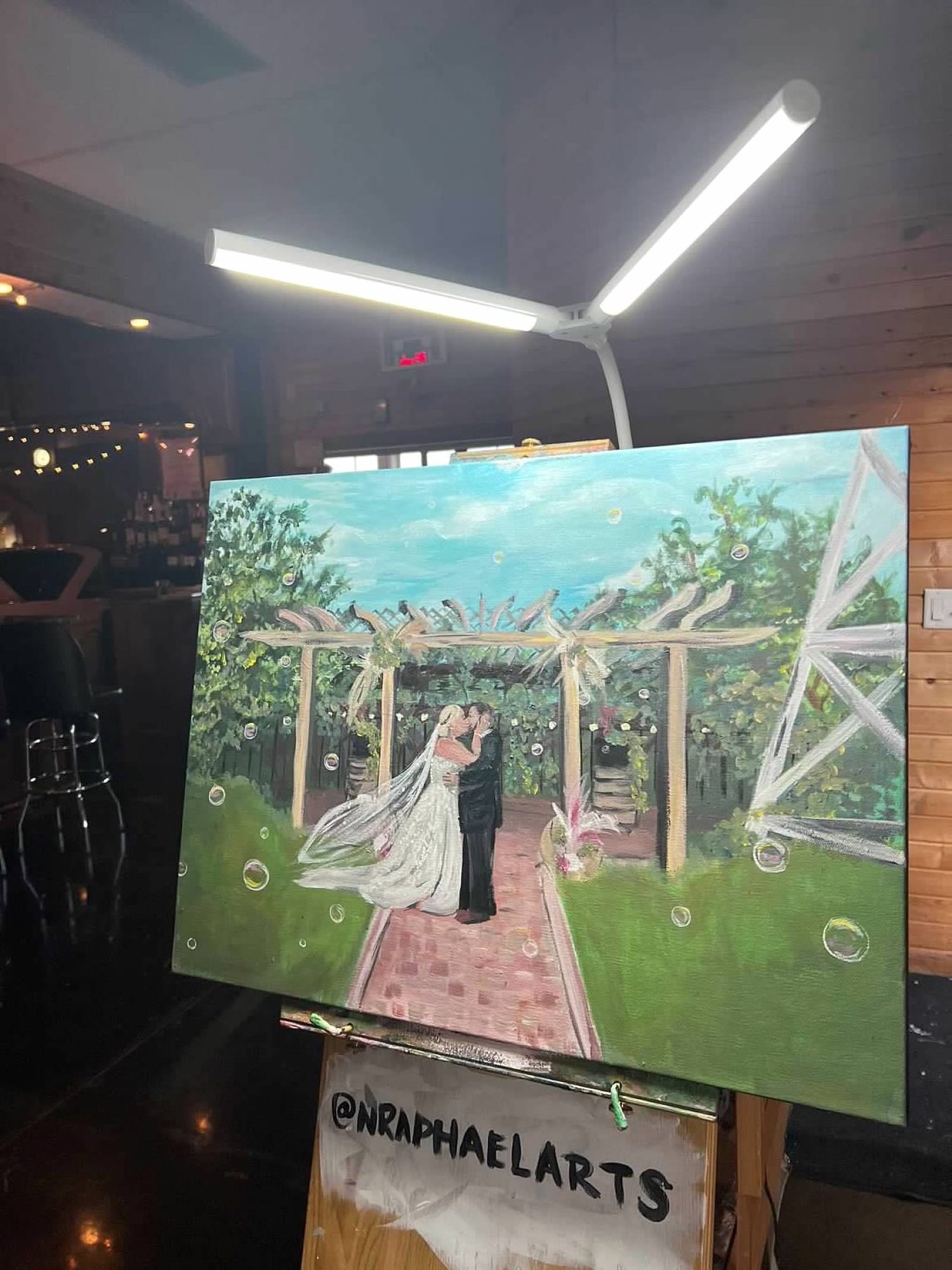 Live canvas painting of a bride and groom under a garden pergola, on an easel at the wedding