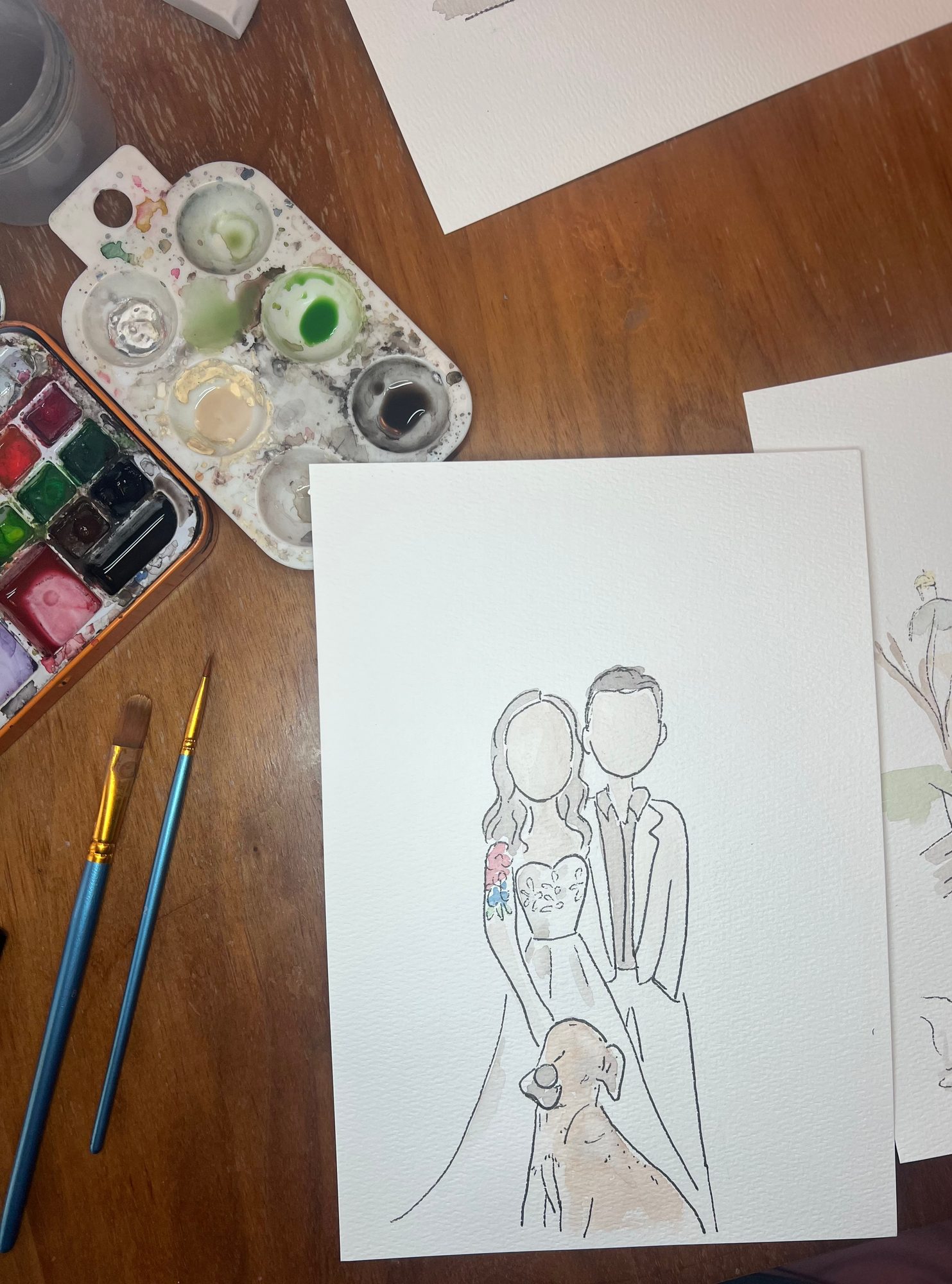 In-progress ink portrait of a bride, groom, and their dog beside a watercolor palette