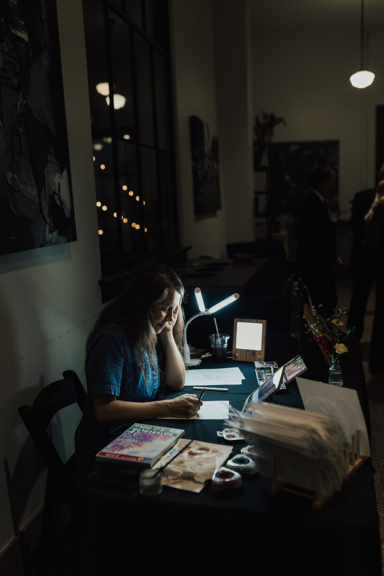 Nicole painting at her lamp-lit table during an evening reception