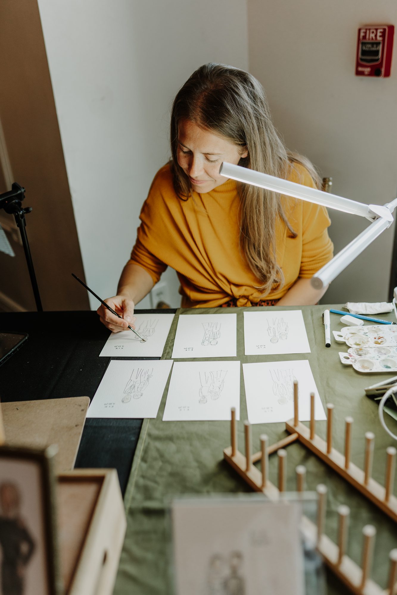 Nicole painting guest portraits at her table, finished minis drying on a rack